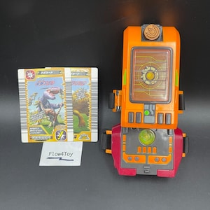 Op de afbeelding: Een oranje en donkerrood plastic polsspeelgoed met een rechthoekig scherm, zwarte bandjes en een cirkelvormig embleem. Twee ruilkaarten met dinosaurusillustraties en Japanse tekst zijn inbegrepen, samen met een Flow4Toy-label.