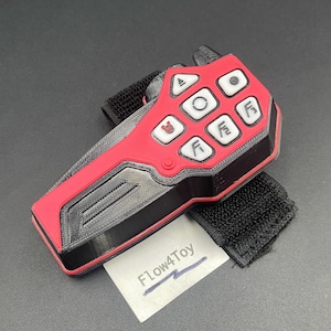 May include: A red and black handheld controller featuring six white buttons with various symbols, including a play icon and stylized letters. The device is attached to a black fabric strap and includes a small white card labeled Flow4Toy.