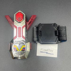May include: A silver and red plastic toy device with a V-shaped emblem featuring yellow, blue, and red stripes, alongside a black rectangular belt buckle component. A small white card with the printed text Flow4Toy is positioned below the buckle.