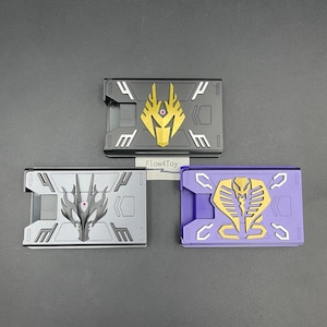 May include: Three rectangular card cases featuring mechanical designs. The top case is black with a gold dragon emblem. The bottom left case is grey with a black dragon emblem. The bottom right case is purple with a gold cobra emblem. Text reads Flow4Toy.