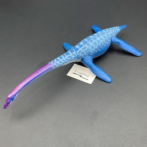 May include: A plastic plesiosaur toy featuring a long neck that transitions from a blue and light blue patterned body to a purple and blue head. The figure has four flippers and a small white tag with the text Flow4Toy printed in black.