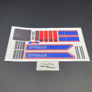 Può includere: Un foglio di adesivi con il testo FIVEMAN in bianco su strisce rettangolari blu e rosse. Il set include varie forme geometriche nere, rosse e blu, strisce e un emblema quadrato grigio scuro. Una piccola etichetta riporta la scritta Flow4Toy.