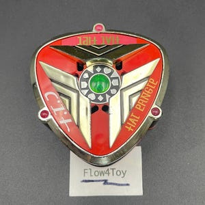 May include: A triangular red and metallic gold toy accessory featuring a central green circular gem, three small red accents at the corners, and text reading "C.I.T." and "HAI PANGTE" in gold lettering. A white card with "Flow4Toy" is visible below.