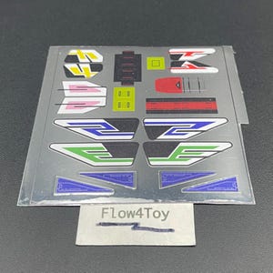 May include: A rectangular sheet of adhesive decals featuring various geometric shapes and lines in yellow, pink, red, blue, green, and black. The sheet includes stylized numbers and abstract patterns. A small white label at the bottom reads Flow4Toy.