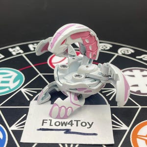 Könnte beinhalten: Eine weiß-rosa Bakugan-Spielfigur in einer teilweise geöffneten, drachenähnlichen Konfiguration mit beweglichen Segmenten und sichtbaren Metallschrauben. Eine kleine weiße Papierkarte mit dem Text Flow4Toy befindet sich vor der Figur.