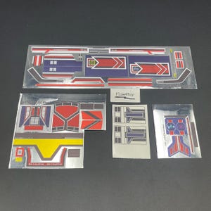 May include: A collection of four metallic sticker sheets featuring geometric patterns in red, blue, yellow, and white. The designs include various shapes, lines, and text such as SKYALPHA and 03-LANDGAMMA. A small white card with the text Flow4Toy is included.