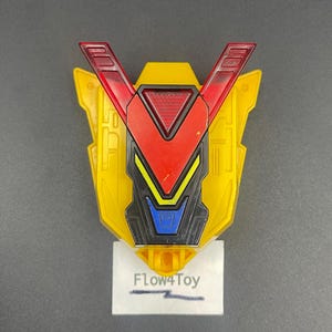 May include: A yellow plastic toy shield with a V-shaped red translucent top section, a central black panel with yellow and blue accents, and a white rectangular base label featuring the printed text Flow4Toy in black ink.