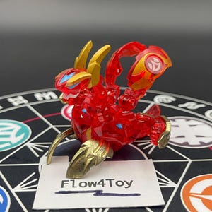 以下が含まれることがあります： 半透明の赤いパーツ、メタリックゴールドの角、脚部の金色のアクセントが特徴的な、赤と金のプラスチック製ドラゴン型アクションフィギュア。背中には円形のエンブレムがあり、Flow4Toyと書かれた白いカードの前に配置されています。