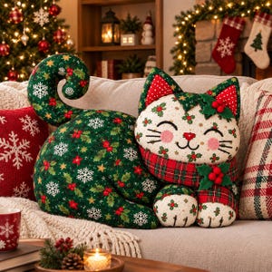 May include: A stuffed cat-shaped cushion featuring a dark green body with a pattern of white snowflakes, red berries, and green holly leaves. The cat has a cream-coloured face with holly prints, a red plaid scarf, and felt holly leaf accents.