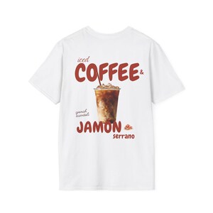 Pode incluir: Uma t-shirt branca de manga curta com um gráfico de um café gelado num copo de plástico transparente. O texto diz iced COFFEE & spanish essentials JAMON serrano, com uma pequena ilustração de uma fatia de presunto ao lado.
