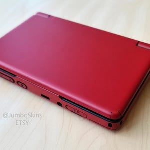 May include: A matte red handheld gaming console with a clamshell design. The device features black trim along the edges, visible buttons on the side, and the text @JumboSkins ETSY printed in small black letters on the lower left corner of the casing.