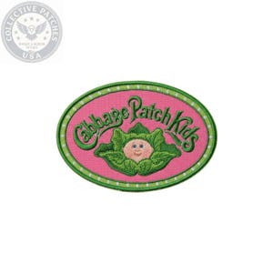 May include: An oval-shaped embroidered patch featuring a pink background with a green border containing white dots. The text Cabbage Patch Kids is embroidered in green above a central illustration of a small doll face nestled in green leaves.