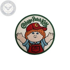 May include: A circular embroidered patch featuring the Cabbage Patch Kids logo in green text above a cartoon character with orange hair, a red cap with a yellow C, blue eyes, and red overalls over a blue shirt, all bordered by a dark green edge.