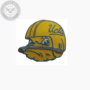 May include: An embroidered patch featuring a yellow football helmet with a blue outline and the text UCLA in blue script. The helmet design includes a stylized mascot face with a blue face mask and blue detailing on a white background.
