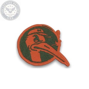 Puede incluir: Un parche bordado circular con fondo verde oscuro y borde naranja. El diseño muestra un pájaro naranja estilizado que lleva una gorra de béisbol, con un pico largo que se extiende más allá del borde circular hacia el lado derecho.