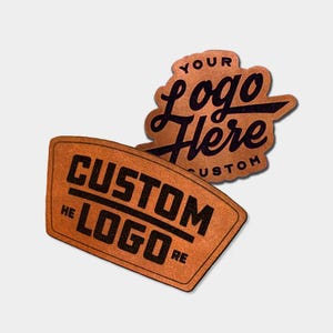 May include: Two tan leather patches with black text. One patch is rectangular with the text CUSTOM LOGO HERE. The other patch is a cloud-like shape with the text YOUR LOGO HERE CUSTOM. Both patches feature a textured surface and bold lettering.