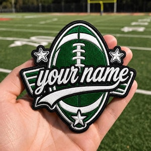 Könnte beinhalten: Ein gestickter Aufnäher in Form eines Footballs in Dunkelgrün und Weiß. Das Design zeigt den Text "your name" in weißer Schreibschrift in der Mitte, akzentuiert durch drei weiße Sterne und einen schwarzen Rand, was ein sportliches Emblem ergibt.