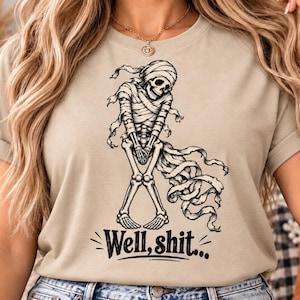 Könnte beinhalten: Ein beiges Kurzarm-T-Shirt mit einer schwarzen grafischen Illustration eines Skeletts, das in Mumienbinden gewickelt ist. Das Skelett steht mit gekreuzten Beinen und gefalteten Händen. Unter der Grafik steht der Text Well, shit...