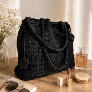 May include: A black crochet tote bag featuring a textured stitch pattern and two matching shoulder straps. A small, decorative crocheted flower charm hangs from the side of the bag. The bag is constructed from dark yarn with a consistent weave.