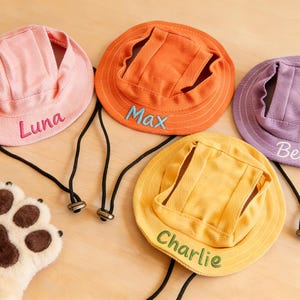 May include: Four pet bucket hats in pink, orange, yellow, and purple, each featuring an adjustable black chin strap and embroidered names: Luna, Max, Charlie, and Bella. A plush, cream-coloured animal paw glove with brown pads is positioned nearby.