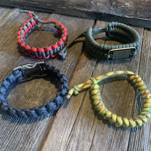 May include: Four paracord bracelets: one red and gray braided cord, one olive green cord with a rectangular metal tag featuring an American flag engraving, one solid dark gray cord with a silver-tone metal buckle, and one yellow, red, and green cord.