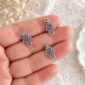 Puede incluir: Tres pequeños dijes de metal plateado con forma de mano de Hamsa. Cada dije presenta un intrincado diseño de filigrana calada con una pequeña anilla circular unida en la parte superior para colgar. Los dijes tienen un acabado texturizado.