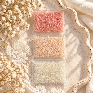 May include: Three rectangular clear plastic pouches filled with small round beads. The top pouch contains pink beads, the middle pouch contains peach-coloured beads, and the bottom pouch contains white beads. The beads have a glossy finish.