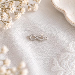 Può includere: Un connettore a forma di simbolo dell'infinito color argento, decorato con una serie di piccole pietre chiare e scintillanti incastonate su tutta la superficie. Il ciondolo presenta un piccolo occhiello circolare a ogni estremità.