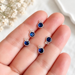 May include: Five small circular charms featuring deep blue faceted stones set in polished silver-toned metal bezels. Each charm includes a small jump ring at the top for attachment, showcasing a consistent design with a vibrant blue gemstone center.