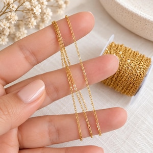 Op de afbeelding: Een fijne goudkleurige metalen ketting met kleine, gelijkmatige ovale schakels. De ketting wordt getoond als een lange, doorlopende streng en als een opgerold stuk rond een kleine witte plastic spoel voor opslag en organisatie.