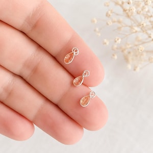 May include: Three small silver charms, each featuring a teardrop-shaped orange gemstone set in a metallic frame with a circular jump ring at the top. The charms are arranged in a vertical line, showcasing the faceted texture of the stones.