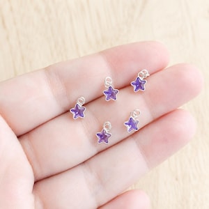 May include: Five small star-shaped charms featuring faceted purple crystals set in silver-toned metal frames. Each charm includes a small circular jump ring at the top for attachment, showcasing a consistent design and vibrant violet color.