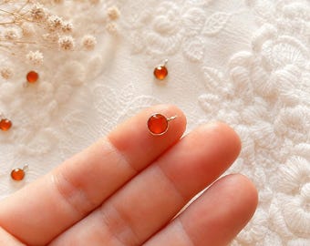 May include: A small, round, orange-red gemstone charm set in a thin silver-toned metal bezel with a tiny jump ring attached at the top. The charm is circular, smooth, and polished, featuring a vibrant, translucent orange-red hue throughout.