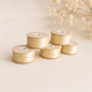 Può includere: Cinque piccole bobine rotonde di filo da cucito color crema chiaro. Ogni bobina presenta un nucleo centrale in plastica bianca ed è avvolta da un filo giallo pallido, liscio e lucido. Le bobine sono disposte in gruppo, mostrando la loro forma cilindrica.