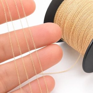 Puede incluir: Una cadena de metal de color dorado con pequeños y delicados eslabones ovalados. La cadena está enrollada alrededor de un carrete de plástico negro, con varios hilos de cadena extendiéndose hacia afuera, mostrando la textura fina y uniforme.