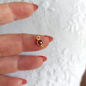 Könnte beinhalten: Ein kleiner Goldanhänger mit einem runden, facettierten, tiefroten Granat-Edelstein in einer Fassung. Der Anhänger verfügt über einen runden Verbindungsring an der Oberseite und drei winzige dekorative Goldkugeln an der Unterseite des Steins.