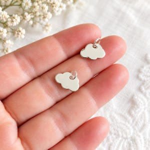 Puede incluir: Dos pequeños dijes en forma de nube de color plateado pulido, cada uno con una superficie lisa y reflectante y una argolla circular unida en la parte superior para colgar. Estos colgantes metálicos de nube tienen bordes redondeados.