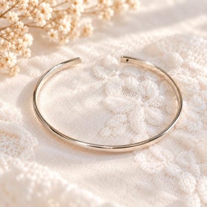 Op de afbeelding: Een gepolijste zilverkleurige metalen manchetarmband met een gladde, ronde band en een open opening. De armband heeft een eenvoudige, cirkelvormige vorm met een klein, donker accentdetail zichtbaar aan een van de open uiteinden.