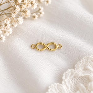 Può includere: Un connettore in metallo color oro a forma di simbolo dell'infinito, caratterizzato da un design a ciclo continuo con un piccolo anello di fissaggio circolare su ciascuna estremità, posizionato orizzontalmente su un tessuto bianco con fiori.