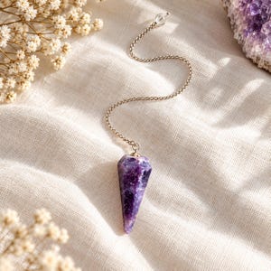Puede incluir: Un péndulo de cristal de amatista morada con forma cónica y facetas, suspendido de una delicada cadena de metal plateado. El cristal presenta tonos violetas intensos con inclusiones blancas naturales y un acabado pulido brillante.