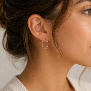 Può includere: Un semplice e sottile orecchino a cerchio in argento indossato sul lobo. L'orecchino è di forma circolare con una finitura metallica lucida e riflettente. Presenta un design essenziale che aderisce all'orecchio con una banda liscia e continua.