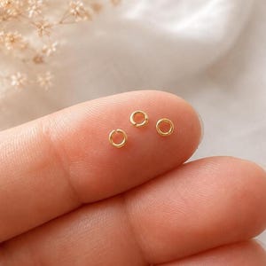 May include: Three small, circular gold jump rings, each featuring a tiny open slit, resting on a surface. These metallic jewellery components are round in shape and possess a polished, reflective gold-toned finish, designed for connecting jewellery pieces.