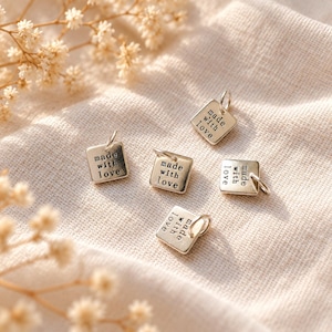 May include: Five small square silver-toned metal charms, each featuring a jump ring at the top corner. The surface of every charm is stamped with the black text "made with love" in a simple, sans-serif font, arranged in three lines.