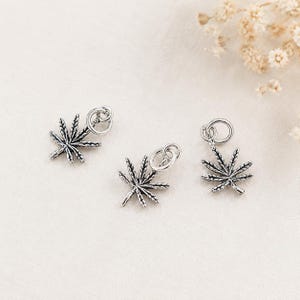 May include: Three silver-toned metal charms shaped like seven-pointed cannabis leaves. Each charm features a detailed, textured surface with dark oxidized accents and includes a small circular jump ring attached to the top for easy hanging.
