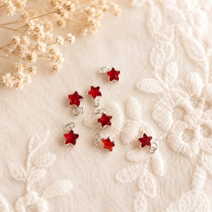 May include: A collection of seven small star-shaped charms, each featuring a vibrant red centre encased in a polished silver-tone metal frame. Every charm includes a small circular jump ring attached to the top point for easy hanging or attachment.