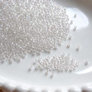 Könnte beinhalten: Eine große Ansammlung kleiner, transparenter, klarer Glas-Saatperlen mit zylindrischer Form. Die winzigen Perlen sind auf einer glatten, weißen, geschwungenen Oberfläche verstreut, mit einer dichten Ansammlung in der Mitte und einigen einzelnen Perlen.