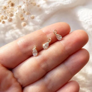 May include: Three small gold-toned charms, each featuring a clear, pear-shaped faceted stone set in a metallic frame with a circular jump ring at the top for attachment. The charms are arranged in a diagonal line, showcasing their reflective surfaces.