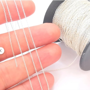 Può includere: Una bobina di sottile catena metallica color argento con piccoli maglie ovali uniformi. La catena è avvolta attorno a un rocchetto di plastica nera e diversi fili della delicata e riflettente catena metallica sono tesi in posizione verticale.