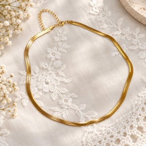 Op de afbeelding: Een goudkleurige visgraatketting met een plat, flexibel schakelontwerp. De ketting is voorzien van een karabijnsluiting en een verstelbare verlengketting met kleine ronde schakels, wat zorgt voor een glad, reflecterend metalen oppervlak.
