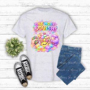 Puede incluir: Una camiseta gris claro con un gráfico colorido de manos apretando varias esferas texturizadas y un pequeño personaje de sol amarillo. El texto sobre el gráfico dice ONE MORE SQUISH! en una fuente de burbujas vibrante y multicolor.